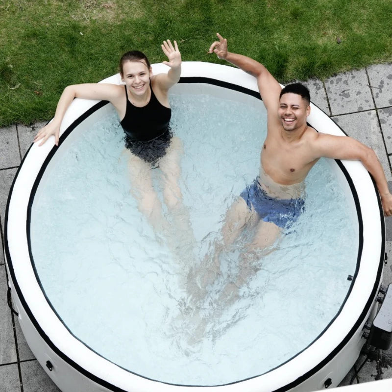 Two people smiling and waving while relaxing in an inflatable hot tub outdoors, enjoying a refreshing soak on a sunny day.